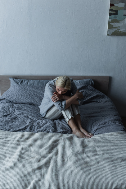 blonde woman hugging knees on bed while feeling pain in stomach during menopause  - Fotoğraf, Görsel