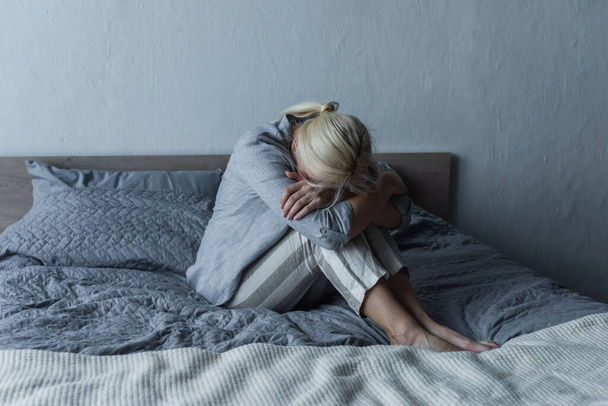 blonde woman covering face and hugging knees on bed while feeling pain in stomach during menopause  - Valokuva, kuva