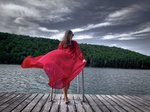 Giovane donna in abito rosso lungo in piedi su un pontone di legno vicino a un lago circondato da una foresta verde - Foto, immagini
