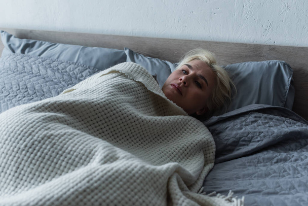 depressed blonde woman with menopause lying under blanket in bed - Foto, afbeelding