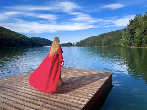 Jeune femme en robe rouge longue debout sur un ponton en bois près d'un lac entouré d'une forêt verte - Photo, image