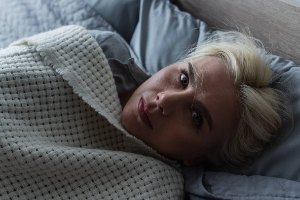 top view of depressed blonde woman with menopause lying under blanket in bed - Foto, Imagem