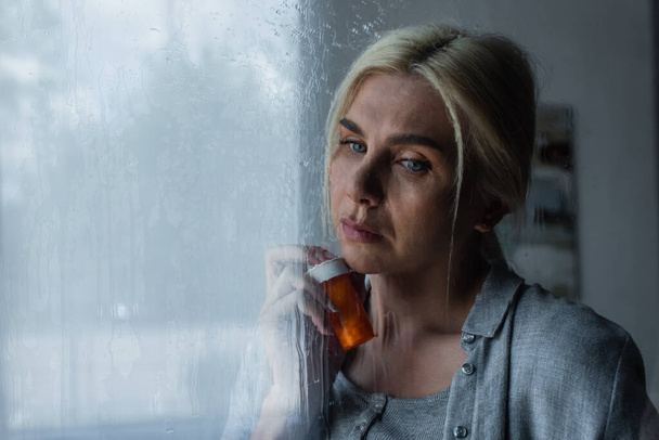 depressieve blonde vrouw houden fles met pillen in de buurt venster met regen druppels - Foto, afbeelding