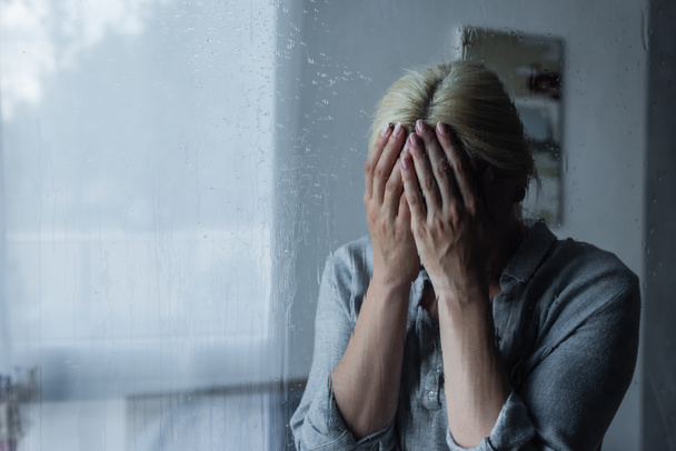 depressed blonde woman covering face behind wet window with rain drops  - Photo, Image