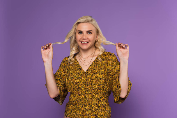 happy and blonde woman in blouse holding curly hair isolated on purple  - Photo, image