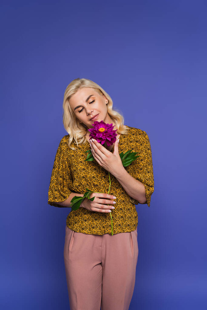 blonde woman in blouse holding flower with green leaves isolated on violet - Fotó, kép