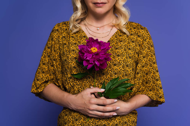cropped view of blonde woman in blouse holding purple flower with green leaves isolated on violet - Фото, изображение