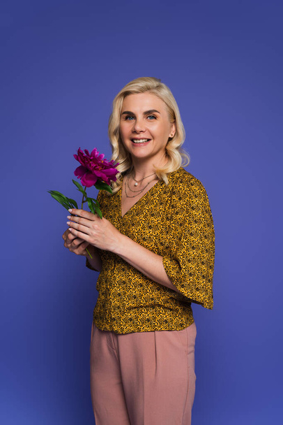 cheerful woman in blouse holding purple flower with green leaves isolated on violet - Foto, Imagem