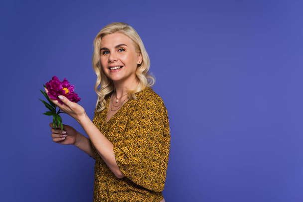positive woman in blouse holding purple flower with green leaves isolated on violet - Foto, immagini