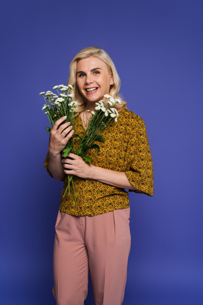 joyful woman in blouse holding bouquet of white flowers with green leaves isolated on violet - Foto, afbeelding