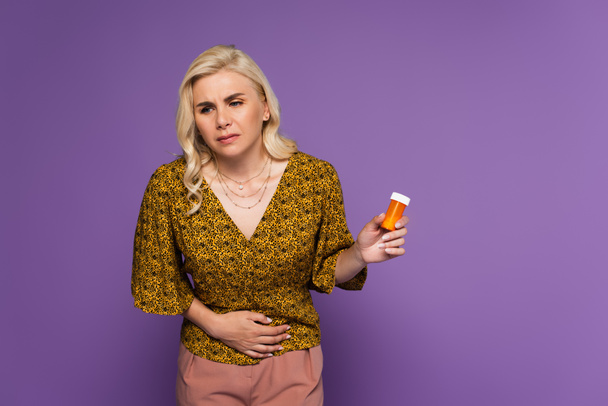 mujer rubia con dolor de estómago sosteniendo botella con pastillas aisladas en púrpura  - Foto, Imagen