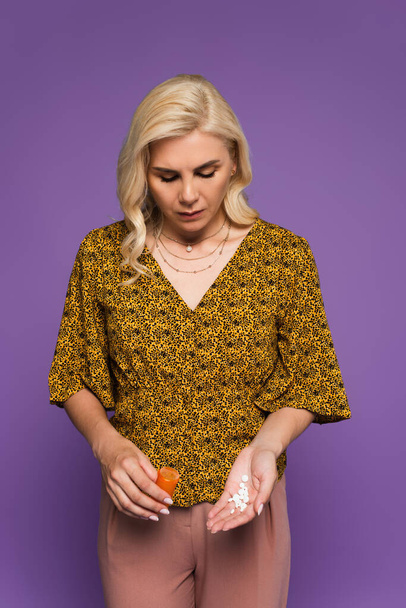 blonde woman with menopause holding bottle and looking at pills isolated on purple  - Photo, Image