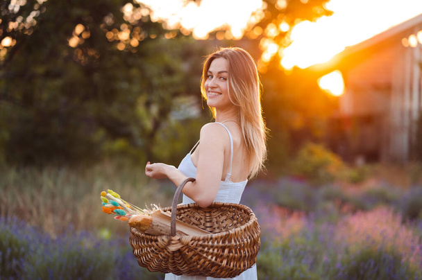 Kaunis valkoihoinen nainen valkoisessa auringonpuvussa kävelee laventeliniityllä kesäiltana kultaiseen aikaan. Kaunis hymy yksinäinen tyttö kylässä kori käsissään - Valokuva, kuva