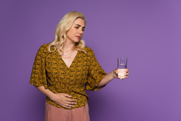 blonde woman with stomach ache holding glass of water isolated on purple  - Zdjęcie, obraz