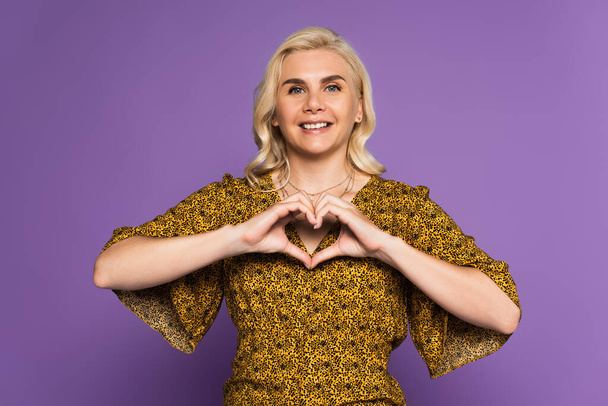 happy blonde woman showing heart sign with hands isolated on purple - Zdjęcie, obraz