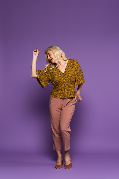 full length of cheerful blonde woman in blouse having menopause and dancing on purple - 写真・画像