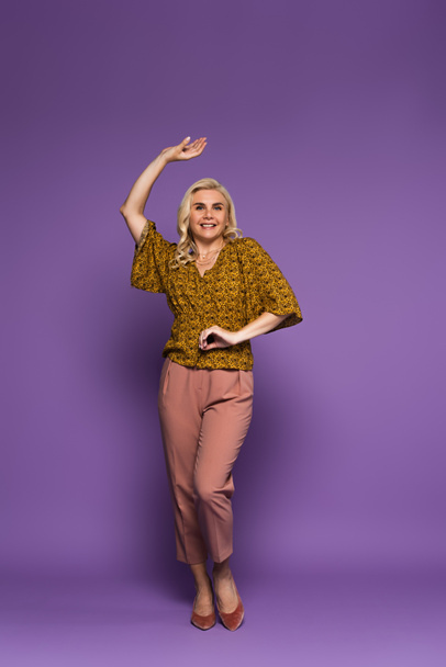 full length of joyful blonde woman in blouse having menopause and dancing on purple - 写真・画像