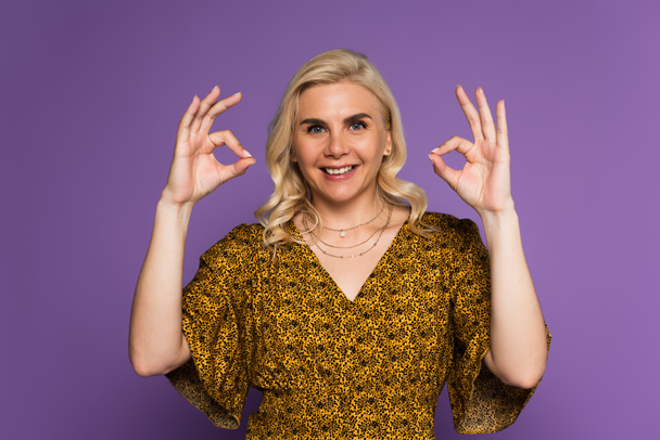 radostná blondýnka s menopauzou s úsměvem a ukazující ok s rukama izolovanými na fialové - Fotografie, Obrázek