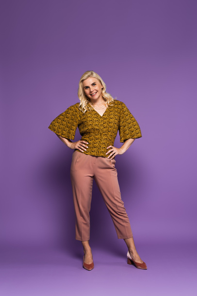 full length of blonde woman with menopause posing with hands on hips and smiling on purple  - Foto, imagen