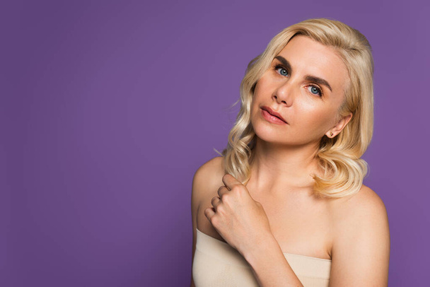 pretty blonde woman with bare shoulders looking at camera isolated on purple - Foto, Imagen