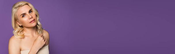 pretty blonde woman with bare shoulders looking at camera isolated on purple, banner - Foto, Imagen