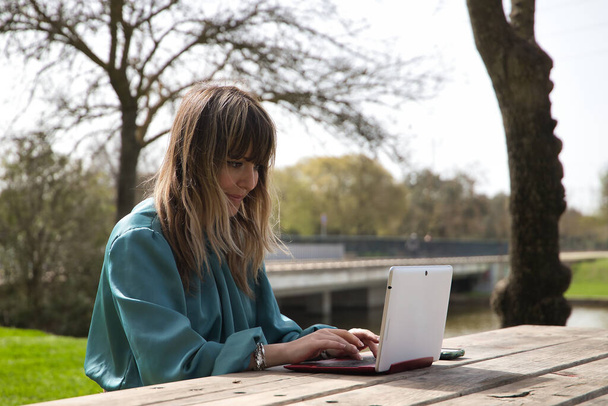 Fiatal, csinos, szőke nő zöld ingben, egy padon ül egy parkban, a laptopjával dolgozik. Technológiai koncepció, tablet, notebook, távmunka, telekommutálás. - Fotó, kép