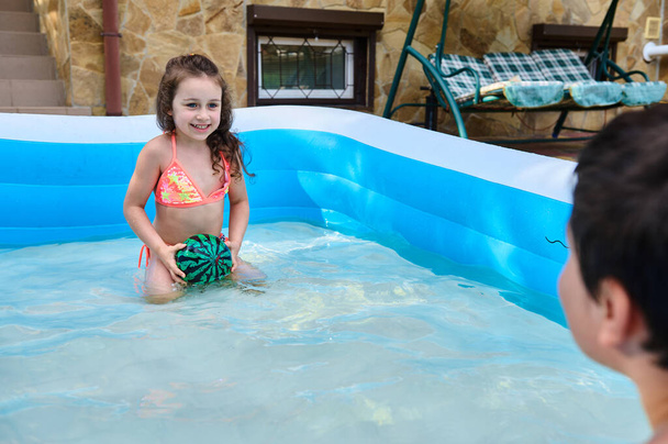 Valkoihoinen lapsi- poika ja tyttö pelaa palloa puhallettavassa uima-altaassa lämpimänä kesäpäivänä aurinkoinen päivä. Sisarukset pitävät hauskaa uima-altaassa. Onnellinen lapsu Kesälomat ja loma käsite - Valokuva, kuva