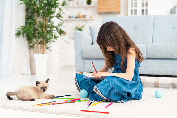 Child girl painting with kitten and sitting on the floor. Little female person drawing and playful kitten next to her at home playing with colorful pencils. High quality photo - Foto, Bild