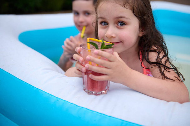 Charming Caucasian kid, cute little girl drinks fresh fruit cocktail and cutely smiles looking at camera while having fun with her older brother in an inflatable swimming pool in the house garden - Fotó, kép