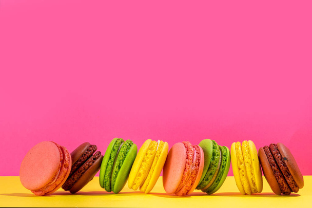 Bright colorful (yellow, pink, green, brown) various flavor macarons sweet cookies on high-colored pink yellow background.  Stack of small french macaron cakes, copy space flatlay  - Фото, изображение