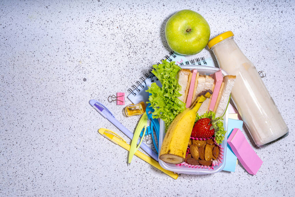 Gezonde schoollunch doos: sandwich, groenten, fruit, noten en yoghurt met schoolkinderen benodigdheden, accessoires en rugzak op zwarte achtergrond platlay kopieerruimte. Terug naar schoolconcept - Foto, afbeelding