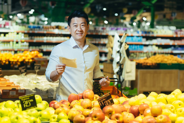 Asian man in supermarket chooses apple fruit, makes purchases looks at shopping list, portrait smiles and looks at camera - Foto, Imagen