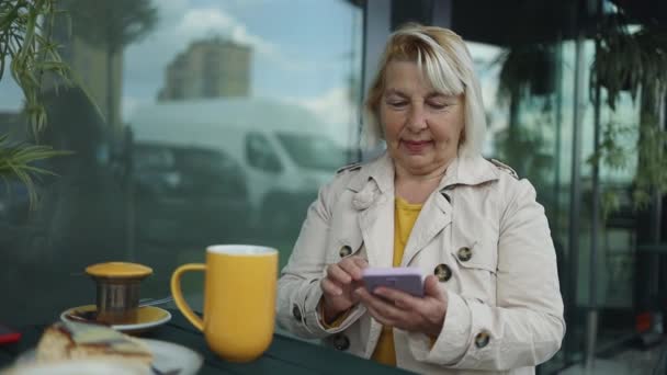 Business freelance 50s blonde woman using smartphone for talking, reading and texting while sitting on table in cafe. Lifestyle smart beautiful women working at coffee shop concepts. - Séquence, vidéo
