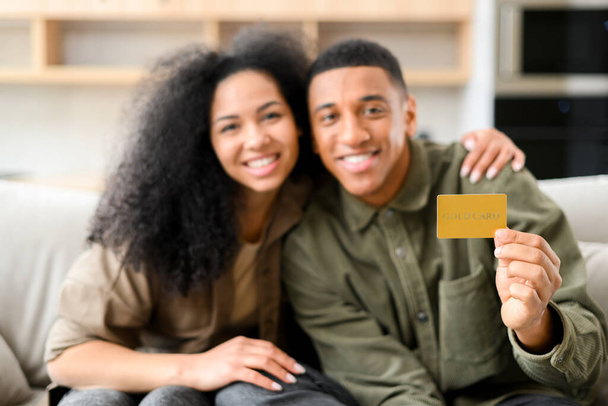 Happy young couple smiling with delight, while sitting at the sofa with credit card and looking at the camera. Happy biracial man showing plastic credit card. Online shopping concept - Foto, afbeelding