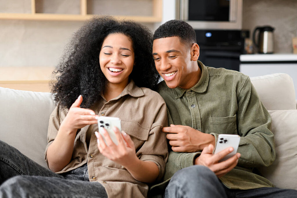 Multiracial couple with smartphone on the couch spending leisure time in social networks, watching videos and discussing something. Black guy bonding to his girlfriend - Φωτογραφία, εικόνα