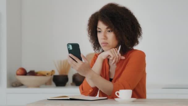 African American μαθήτρια κορίτσι με σγουρά μαλλιά κάθεται στο σπίτι κουζίνα βλέποντας webinar ζωντανά εργαστήριο μάθημα τάξη εικονική συνομιλία smartphone κινητό τηλέφωνο e-learning μελετώντας σημειώσεις γραφής κάνει την εργασία - Πλάνα, βίντεο