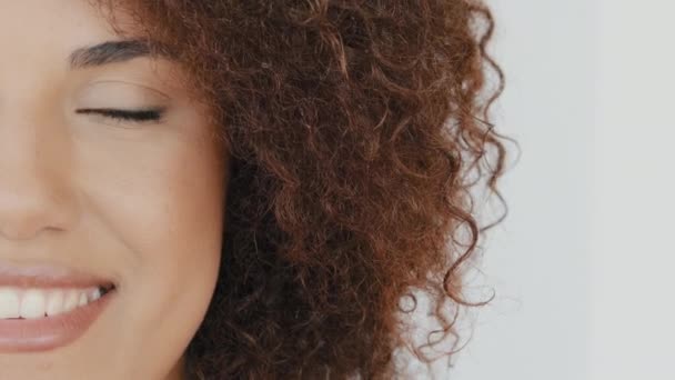 Close up young african american smiling toothy happy curly hair woman face half opening eye and looking to camera with smile. Portrait of pretty female girl with nude natural make up posing indoor - Materiał filmowy, wideo