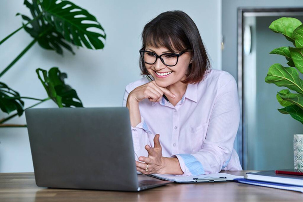 Mature woman using laptop for video call, virtual meeting, conference chat, looking at computer screen talking. Middle aged business female wearing glasses sitting at table in home office - Foto, afbeelding