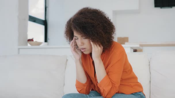 Unhappy African American woman sit on couch at home kitchen living room rubs temple feeling pain closed eyes suffers headache painful feelings chronic migraine hurt worry financial problem break up - Materiaali, video