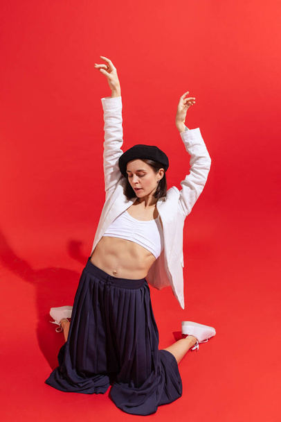 Portrait of stylish young woman posing, dancing isolated over red studio background. Stylish clothes. Concept of beauty, fashion, style, emotions, femininity, facial expression, sales, ad - Foto, afbeelding