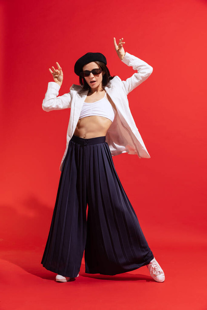 Stylish young woman in black beret and sunglasses posing, dancing isolated over red background. Parisian look. Concept of beauty, fashion, style, emotions, femininity, facial expression, sales, ad - 写真・画像