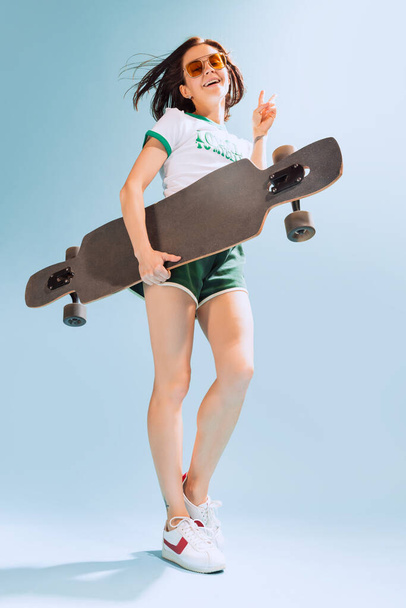 Портрет веселой женщины в непринужденной летней одежде, позирующей со скейтбордом на светло-голубом фоне. Концепция красоты, стиля, эмоций, моды, образа жизни, выражения лица, объявления - Фото, изображение