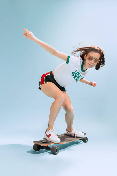 Portrait of happy woman in casual summer outfit posing on skateboard isolated over light blue background. Active hobby. Concept of beauty, style, emotions, fashion, lifestyle, facial expression, ad - Photo, image
