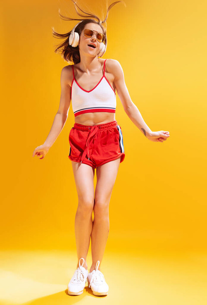 Beautiful young woman in top and red shorts posing, listening to music in headphones isolated over yellow background. Concept of beauty, style, emotions, fashion, lifestyle, facial expression, ad - Photo, image
