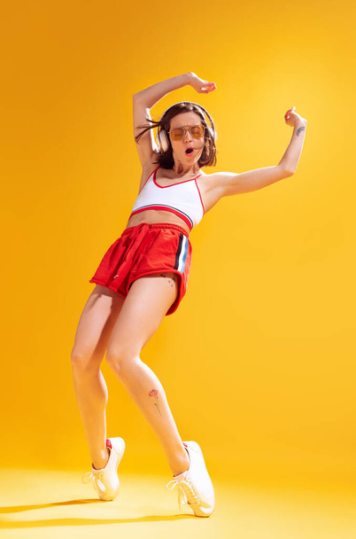 Cheerful young woman in top and red shorts posing, dancing, listening to music in headphones isolated over yellow background. Concept of beauty, emotions, fashion, lifestyle, facial expression, ad - Foto, Imagem