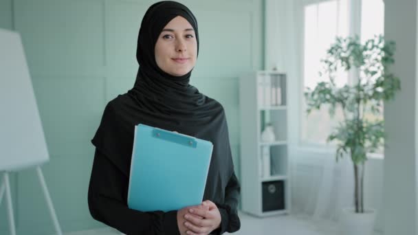 Islamic Indian lady Muslim girl intern standing at modern office coworking space holding folder with documents. Arabic female office manager boss leader worker businesswoman in hijab looking to camera - Metraje, vídeo
