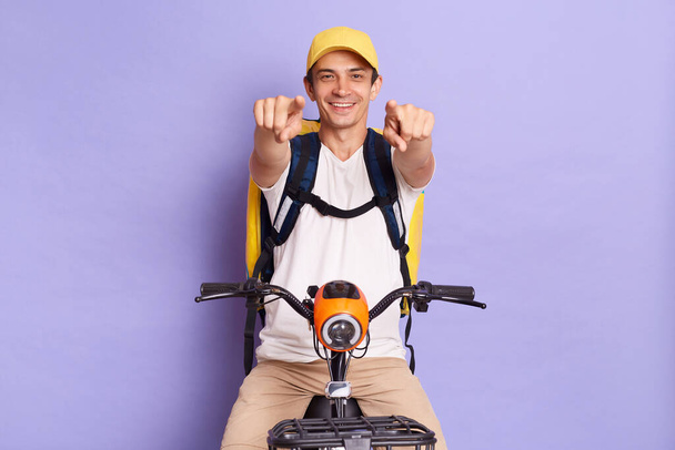 Indoor shot of young satisfied delivery man of takeaway food on electric posing isolated over purple background, pointing at you with happy facial expression. - Fotoğraf, Görsel