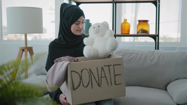 Arabian Islamic Muslim female in black hijab kind woman volunteer at home sitting on couch packing donations box with clothes and toys for kids putting teddy bear in parcel war donating volunteering - Filmagem, Vídeo