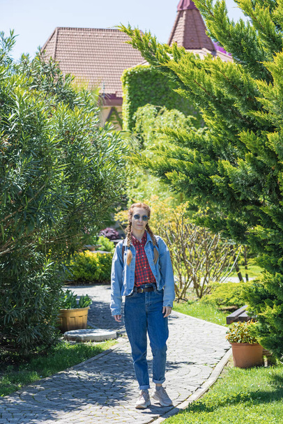 A smiling woman in denim outfit walks along a paved park path passing between lawns planted with dense trees and bushes on a sunny spring day - Photo, image