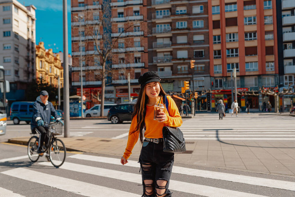 Счастливая азиатская туристка, переходящая пешеходный переход во время осмотра достопримечательностей европейского города, выпивая шоколадный молочный коктейль. Женщина, гуляющая в Хихоне, Астурия, Испания. - Фото, изображение